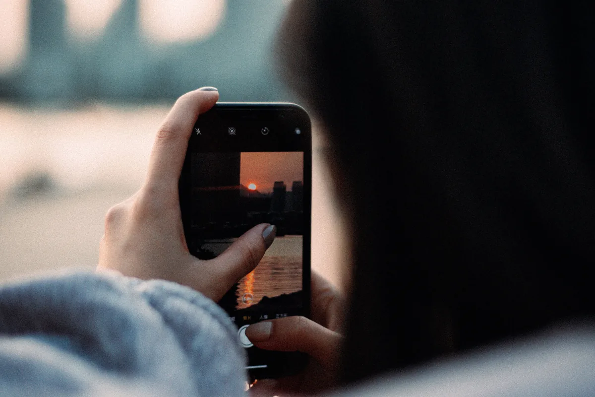 Person fotografiert mit dem Smartphone einen Sonnenuntergang &uuml;ber einer Stadt